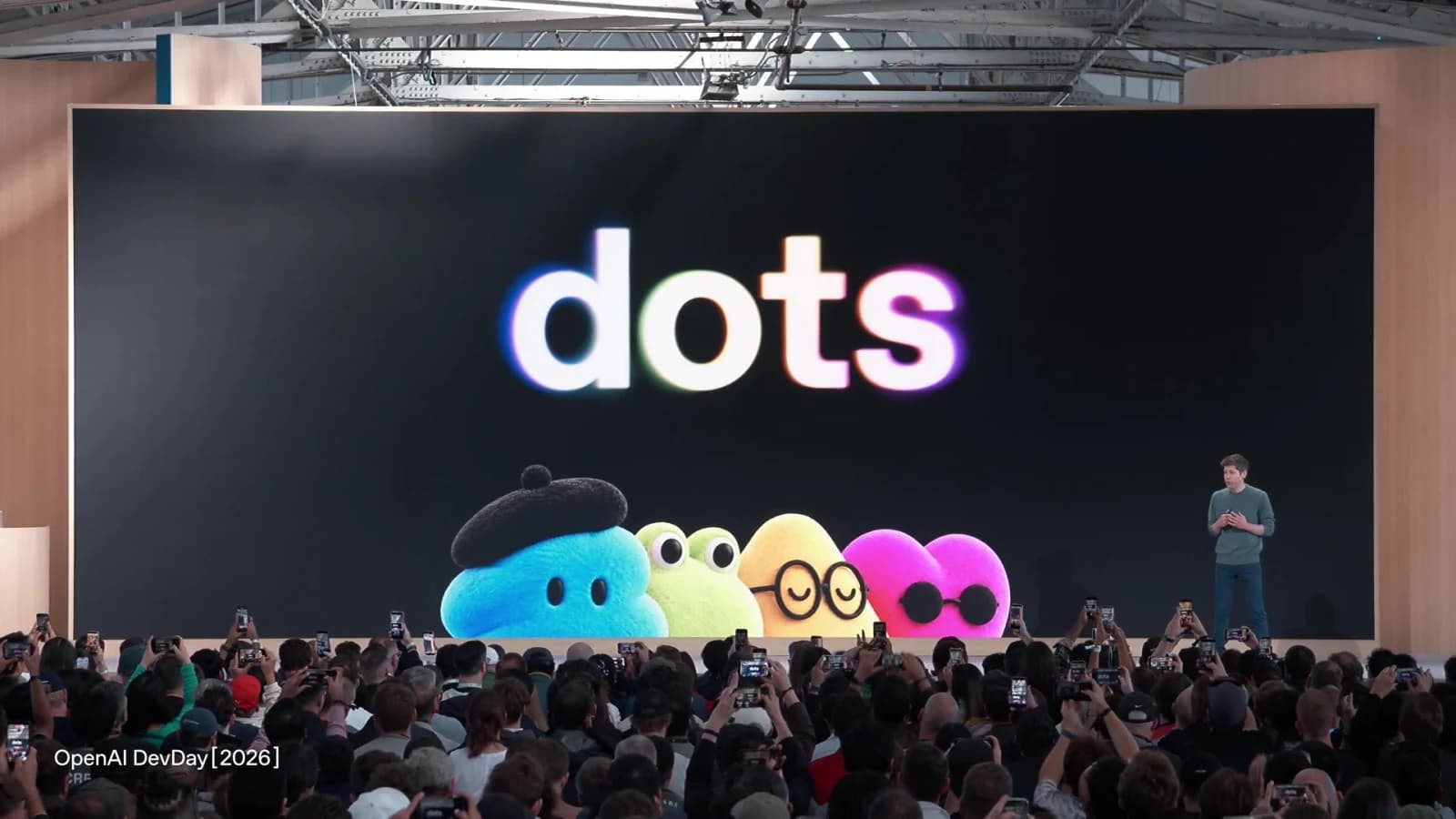 Sam Altman présente les dots sur la scène de la keynote OpenAI DevDay 2026 : le mot dots en lettres lumineuses et quatre personnages en peluche, devant un public qui filme avec ses téléphones. Source : OpenAI, keynote DevDay 2026